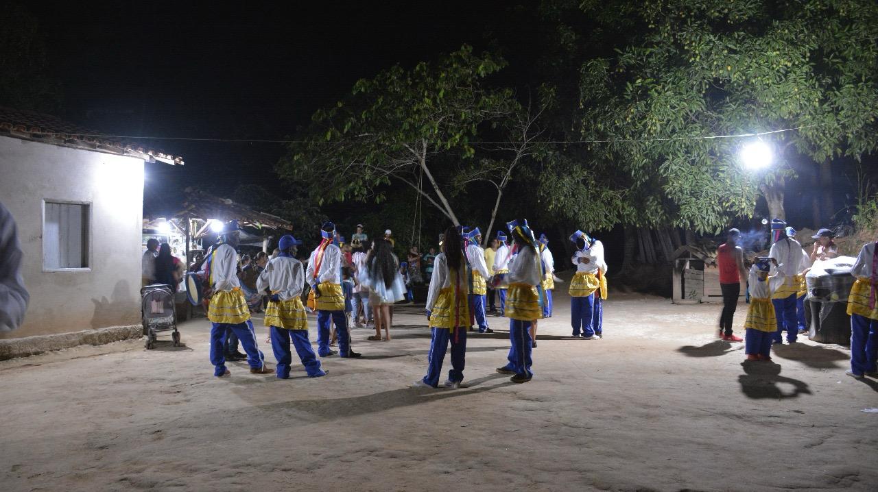 A imagem retrata um grupo de pessoas participando do congado. Elas estão reunidas em um terreiro, com árvores de fundo.