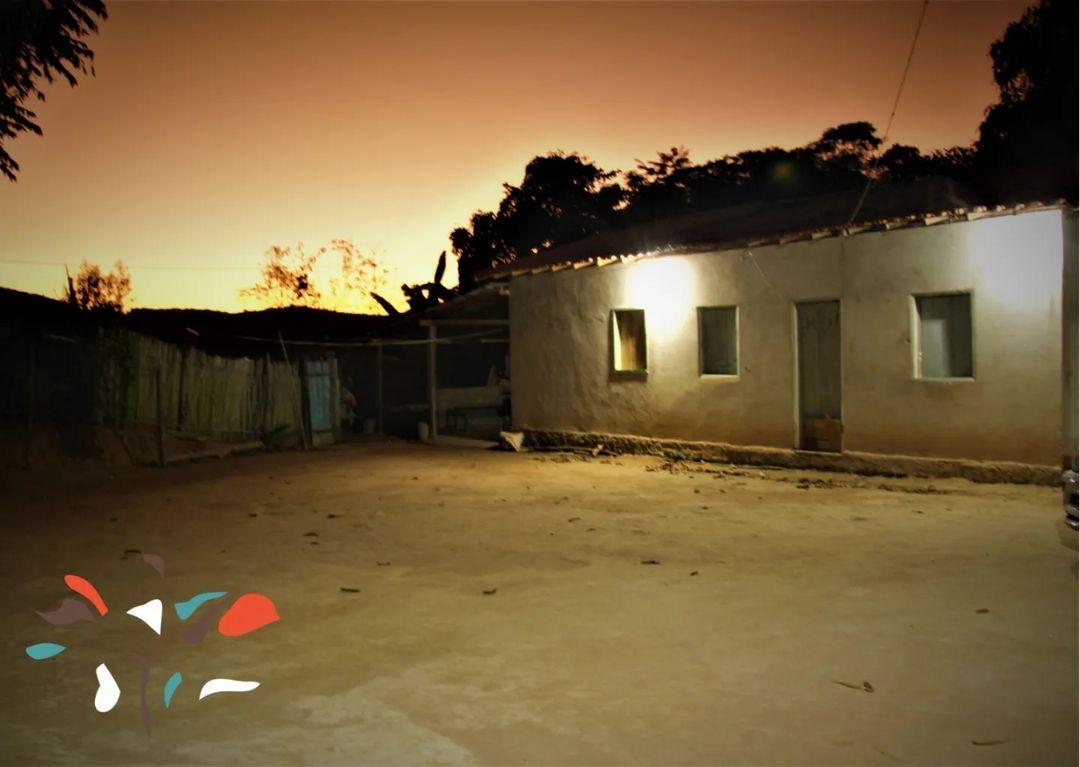 Essa imagem retrata o entardecer no Quilombo dos Bernados, casa simples com paredes brancas e telhado de telhas cerâmicas, iluminada por luzes externas ao entardecer.  O pátio em frente à casa está vazio, cercado por uma cerca de madeira. Ao fundo, a silhueta de árvores e arbustos contra um céu alaranjado.