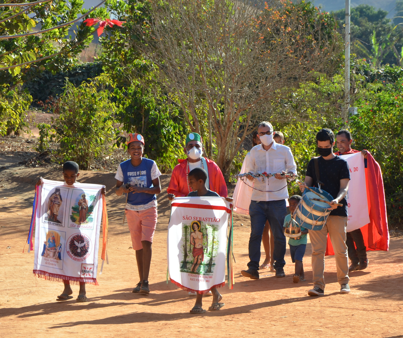 A imagem retrata um grupo de pessoas participando de uma procissão do congado. Elas caminham ao ar livre em um caminho de terra, com vegetação e árvores dos dois lados.