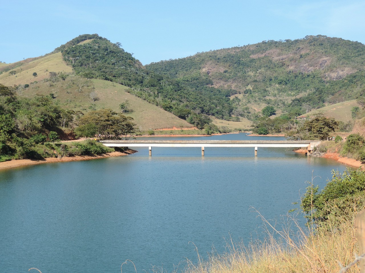 Essa imagem retrata a represa do emboque, uma vista cênica de um reservatório azul calmo cercado por colinas verdes sob um céu claro