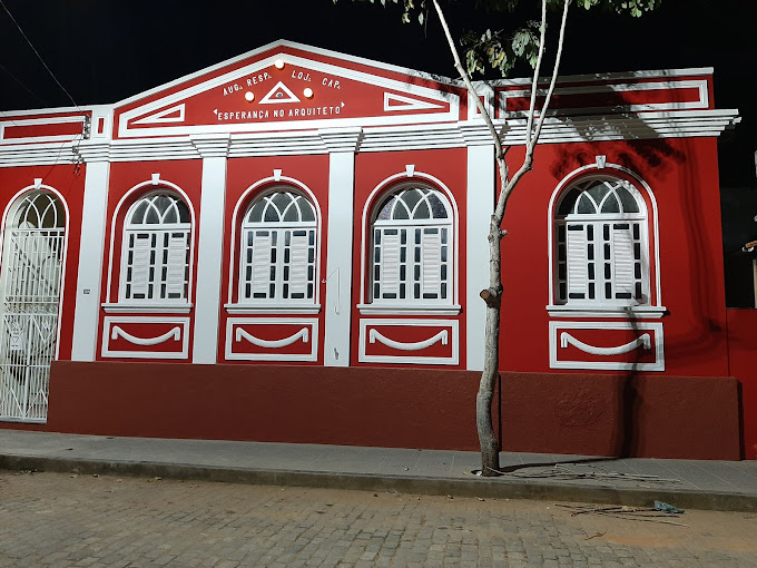 Essa imagem retrata um prédio vermelho vibrante com detalhes em branco, seguindo um estilo arquitetônico clássico. Há seis grandes janelas com molduras brancas e três portas de entrada, também emolduradas em branco. Acima da porta central, há uma placa que diz “LOJA ESPERANÇA DO ARQUITETO”.