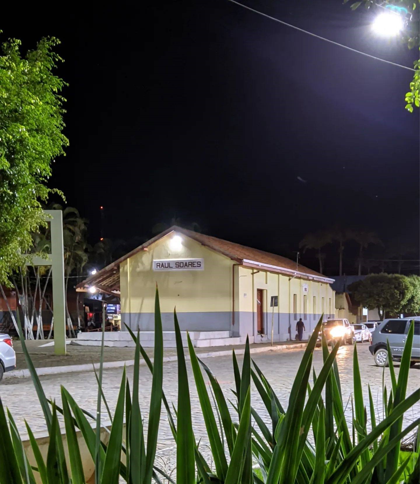 Essa imagem retrata a praça da estação, prédio à noite com a placa “RAUL SOARES” iluminada por luzes de rua, com carros estacionados ao lado e folhagem em primeiro plano.