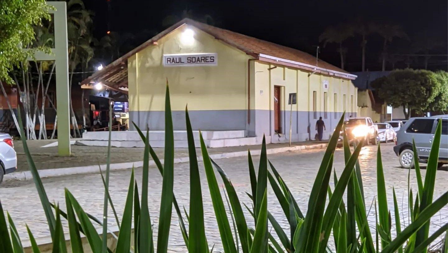 Essa imagem retrata a praça da estação, prédio à noite com a placa “RAUL SOARES” iluminada por luzes de rua, com carros estacionados ao lado e folhagem em primeiro plano.