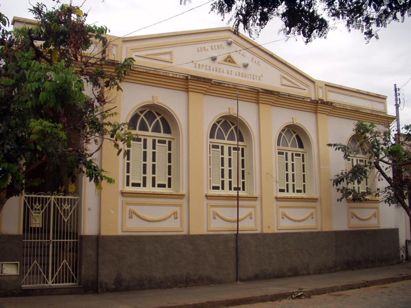 Essa imagem retrata um prédio, seguindo um estilo arquitetônico clássico. Há seis grandes janelas com molduras brancas e três portas de entrada, também emolduradas em branco. Acima da porta central, há uma placa que diz “LOJA ESPERANÇA DO ARQUITETO”.