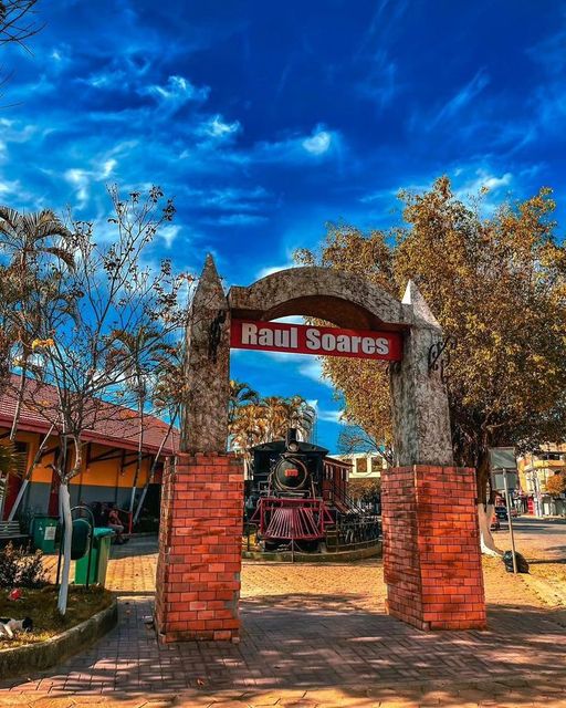 Foto da maria fumaça praca da estacao de com uma placa escrita 'Raul Soares'. A entrada é composta por duas colunas de tijolos com um arco de pedra no topo. Ao fundo, há uma locomotiva antiga e árvores, com um céu azul vibrante e algumas nuvens.
